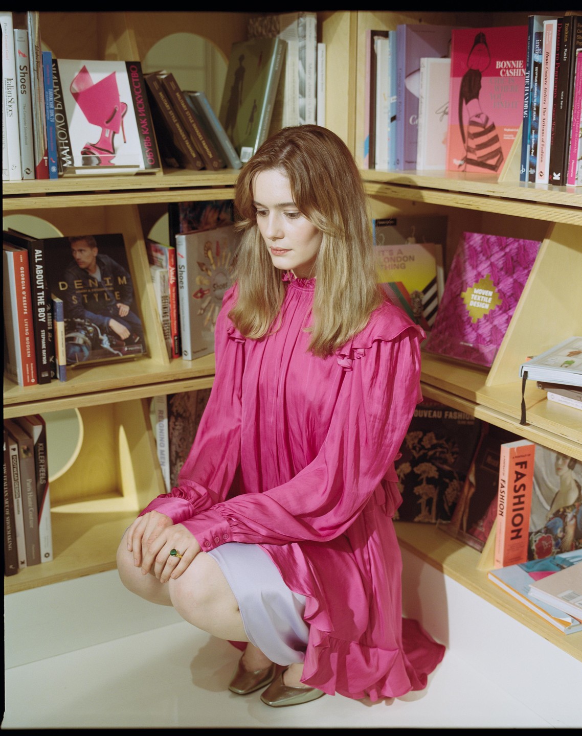 A woman in a pink dress crouches in front of a wooden bookshelf filled with books.