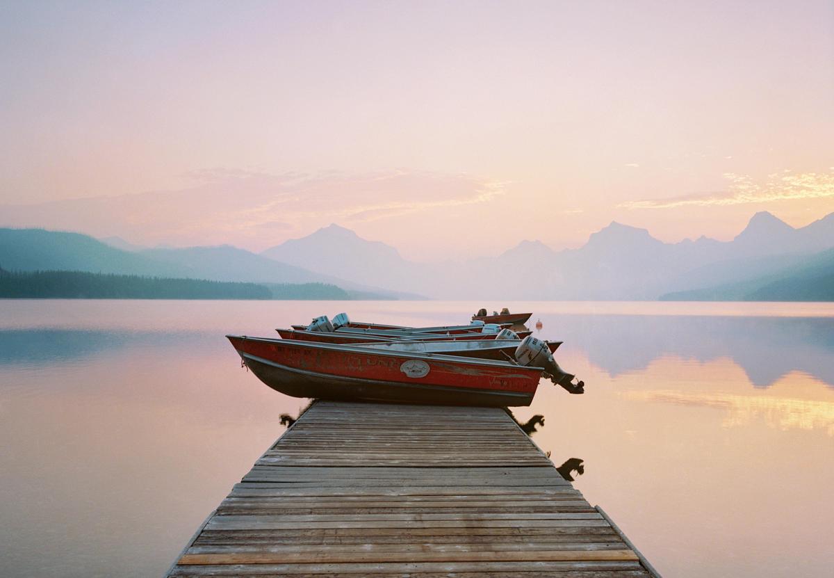 Several red boats are docked on a wooden pier extending into a calm lake with mountains in the background under a pastel sky.