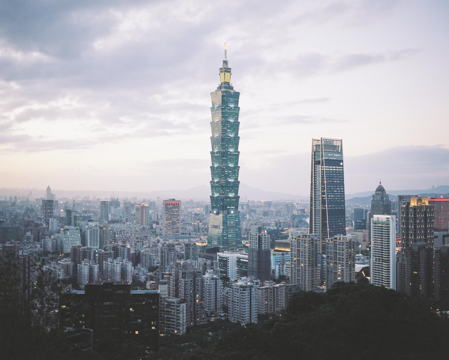 A tall skyscraper with illuminated windows stands prominently among other buildings in a city at dusk.
