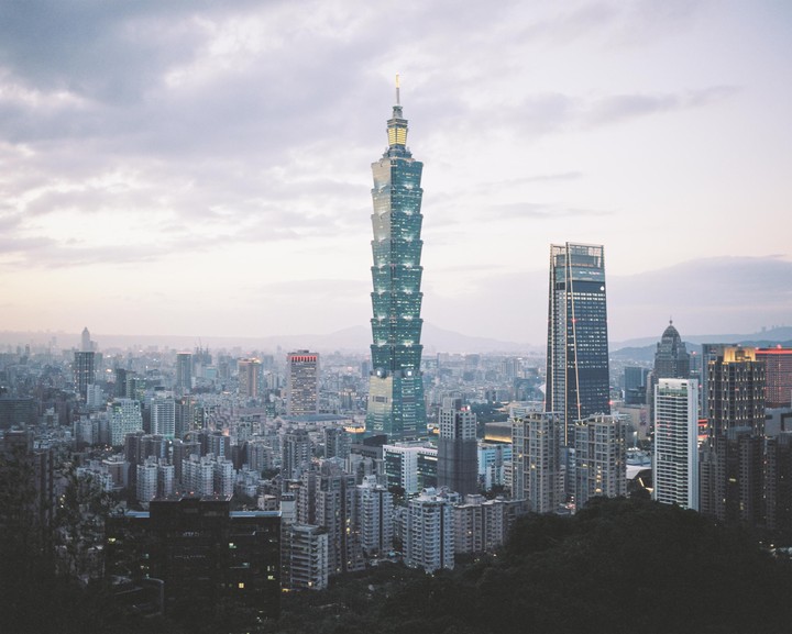 A tall skyscraper with illuminated windows stands prominently among other buildings in a city at dusk.