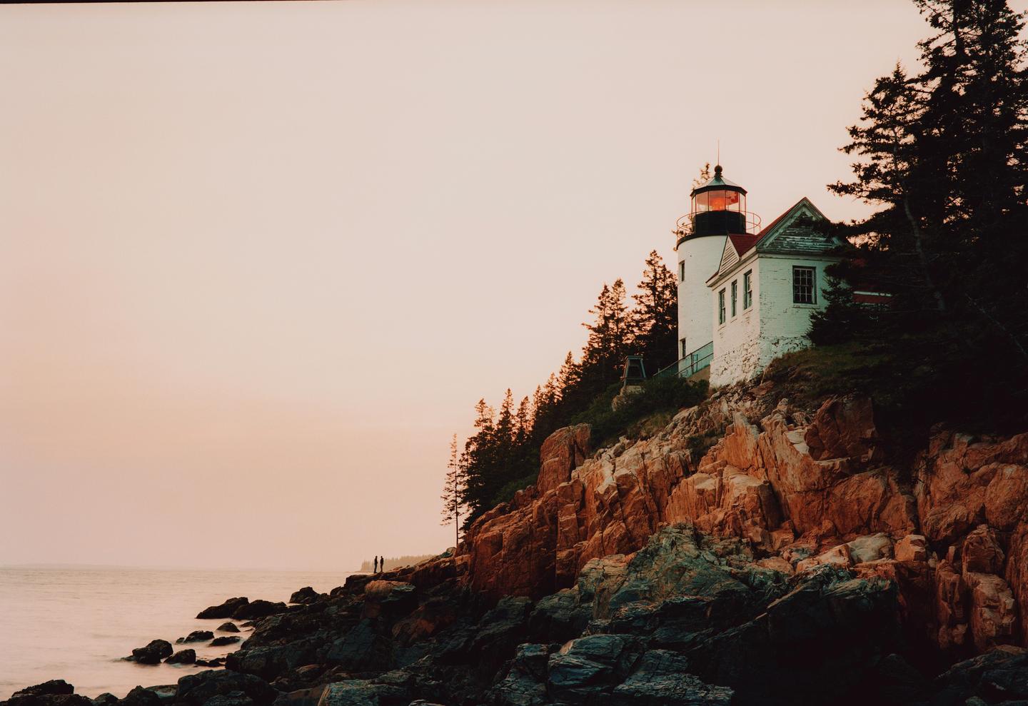 A white lighthouse with a red roof stands on a rocky cliff overlooking the ocean at dusk.