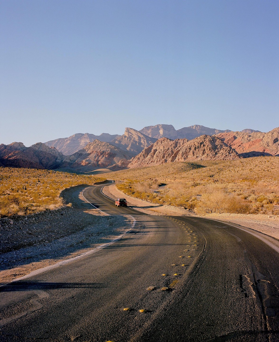 A red car drives on a winding road through a desert landscape with mountains in the background.