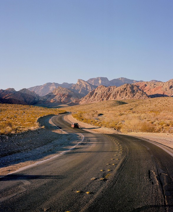 A red car drives on a winding road through a desert landscape with mountains in the background.