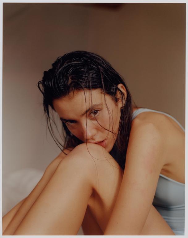 A woman with wet hair is sitting with her knees pulled up to her chest, looking directly at the viewer.