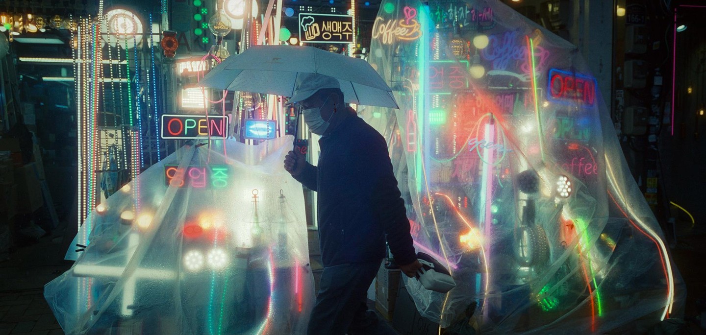 A man with an umbrella and face mask walks past a shop with many neon signs and lights.