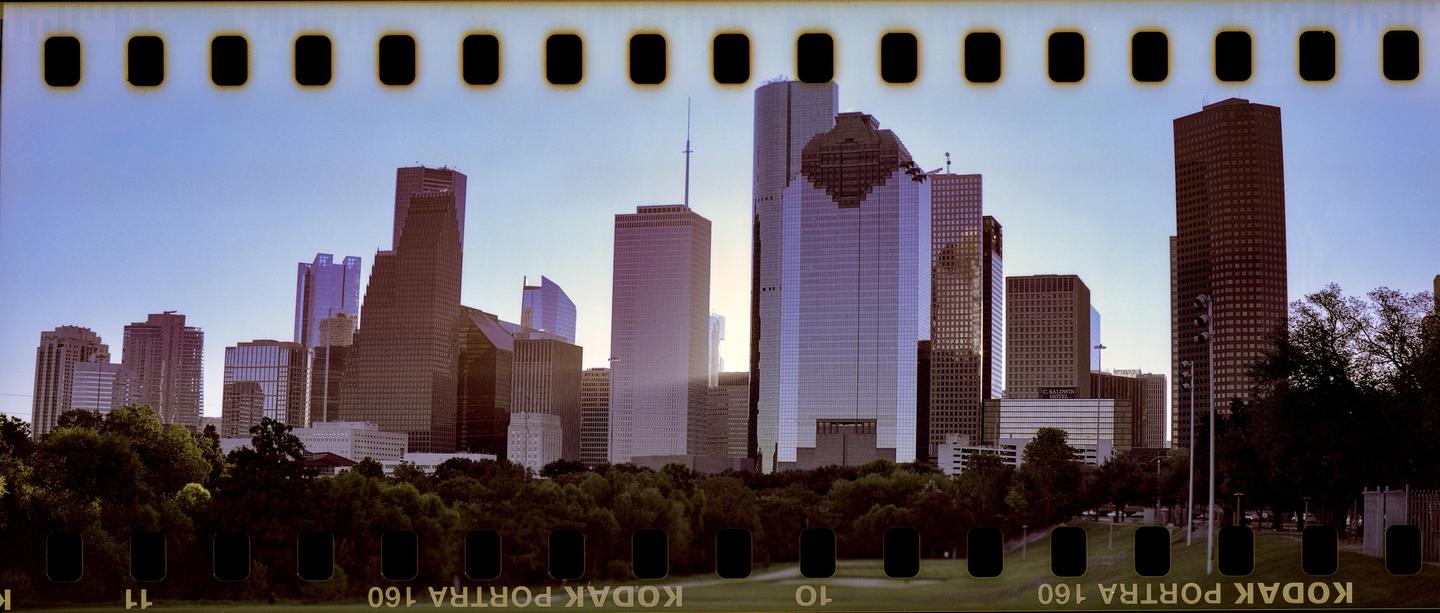 A panoramic view of a city skyline with numerous skyscrapers rising above a line of green trees under a clear sky.