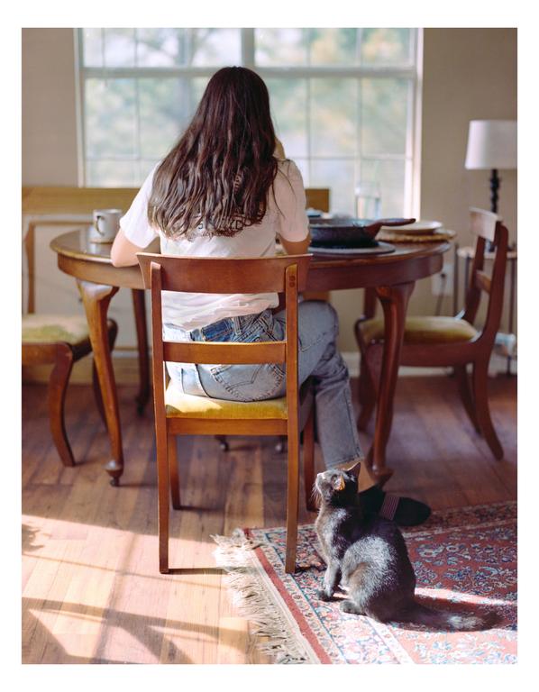 A woman sits at a wooden table with a cat on the floor looking up at her.