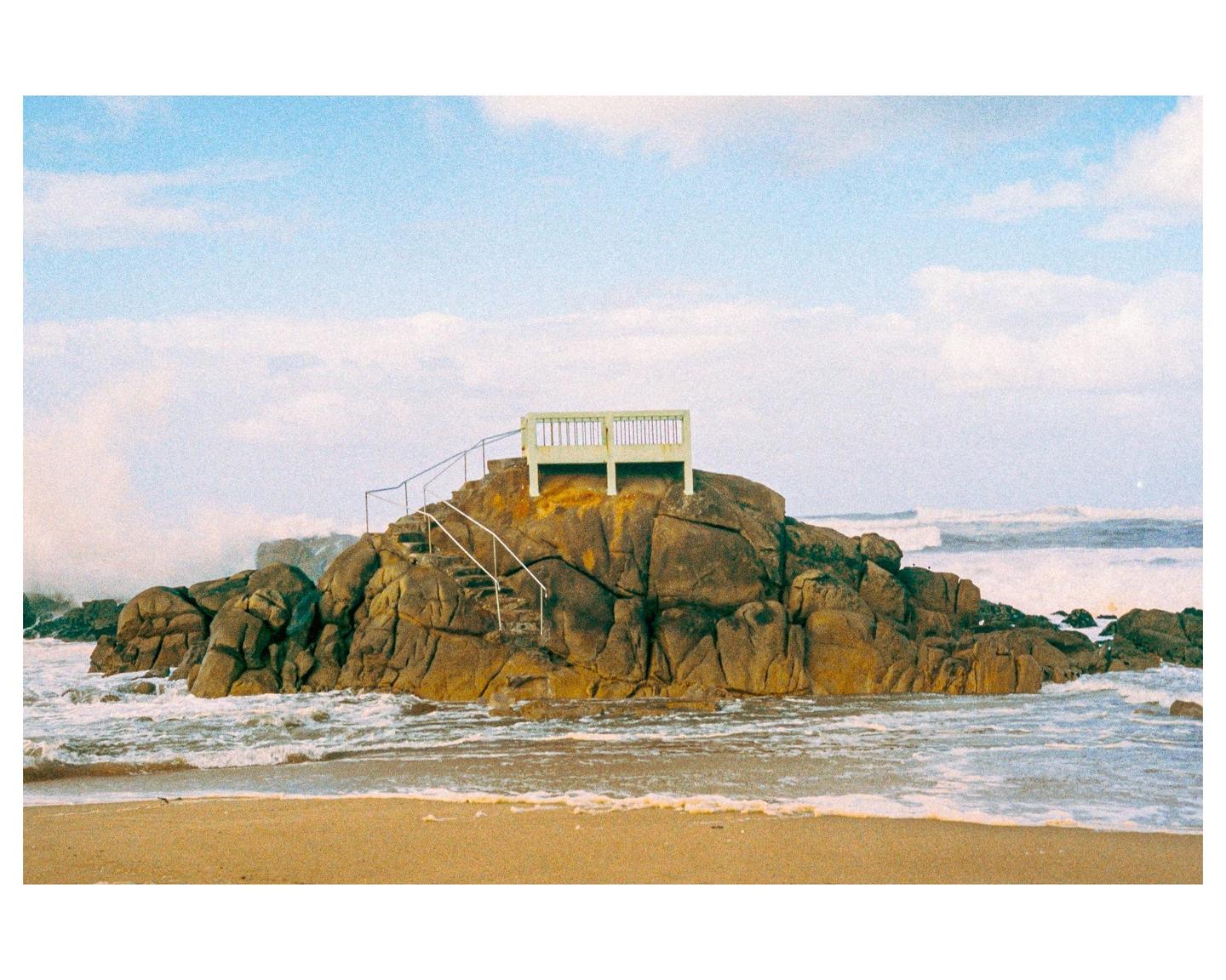 A small wooden structure with stairs sits atop a rocky outcrop on a sandy beach with waves crashing around it.