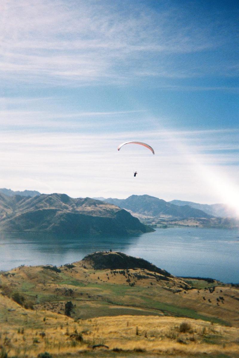A paraglider flies over a lake surrounded by mountains and hills.