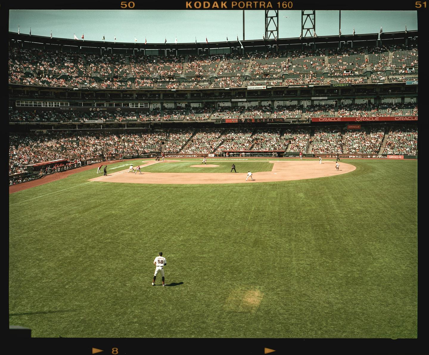 A baseball player stands on a green field with a stadium full of spectators in the background.