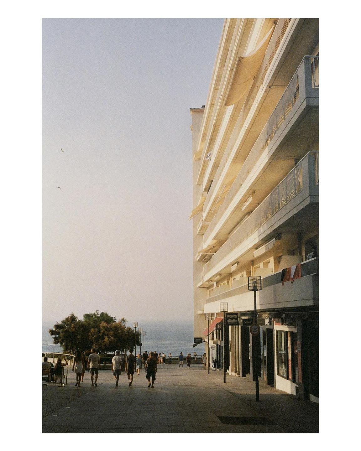 A street with people walking along a tall building and the ocean in the background during golden hour.