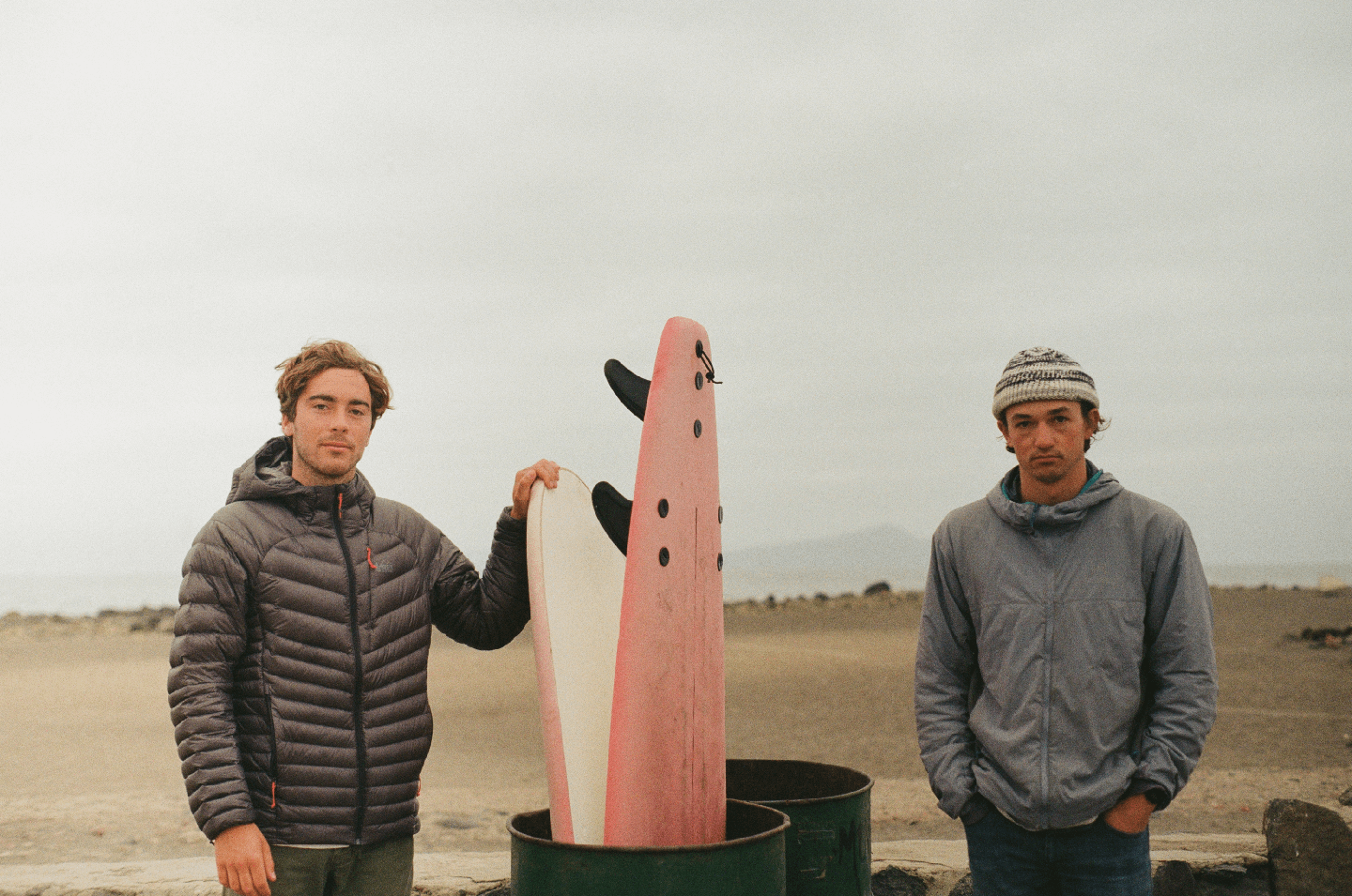 Two men stand on a beach with surfboards in barrels.