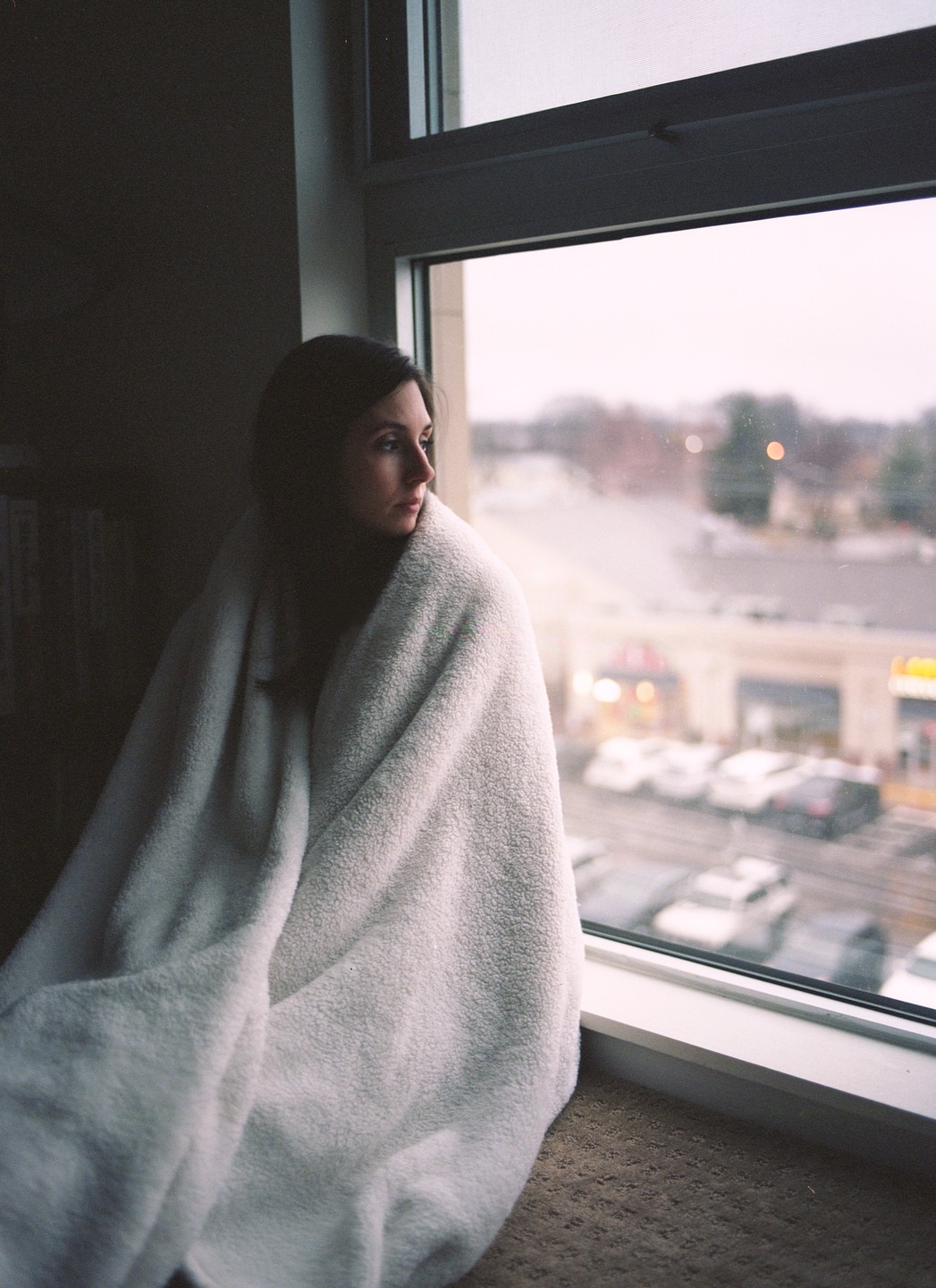 A woman wrapped in a white blanket looks out a window at a parking lot.