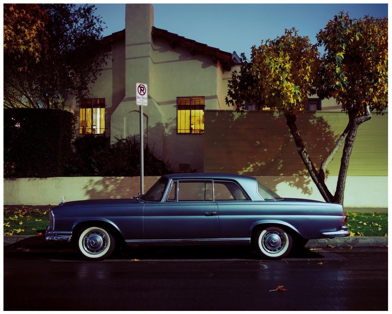 A blue vintage car is parked on a wet street next to a house with illuminated windows and trees with autumn foliage.