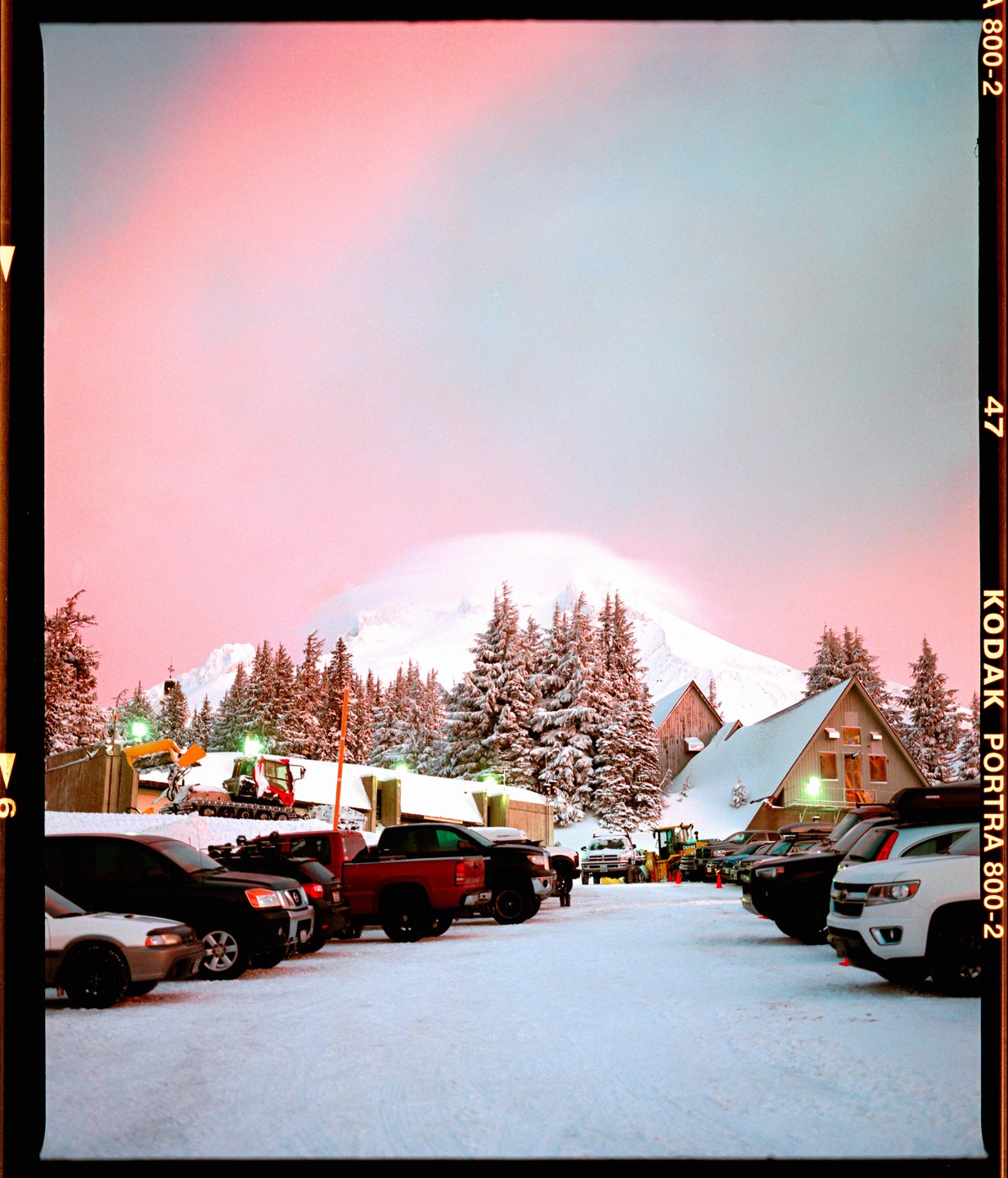 A parking lot full of vehicles is covered in snow with a mountain and trees in the background under a pink and blue sky.