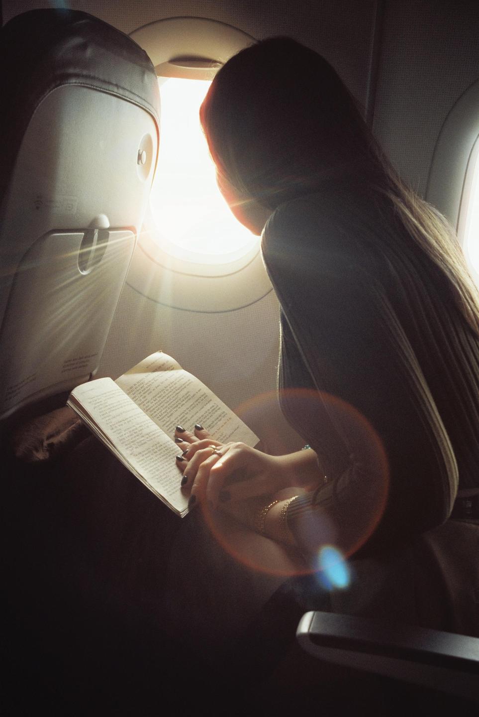A woman reads a book while sitting in an airplane seat next to a window with bright light coming through.