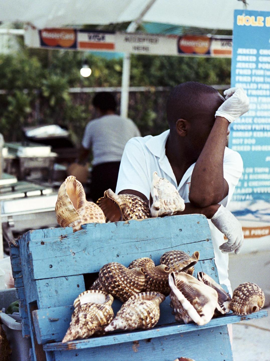 A man in a white shirt and gloves rests his head on his arm next to a blue wooden crate filled with seashells.