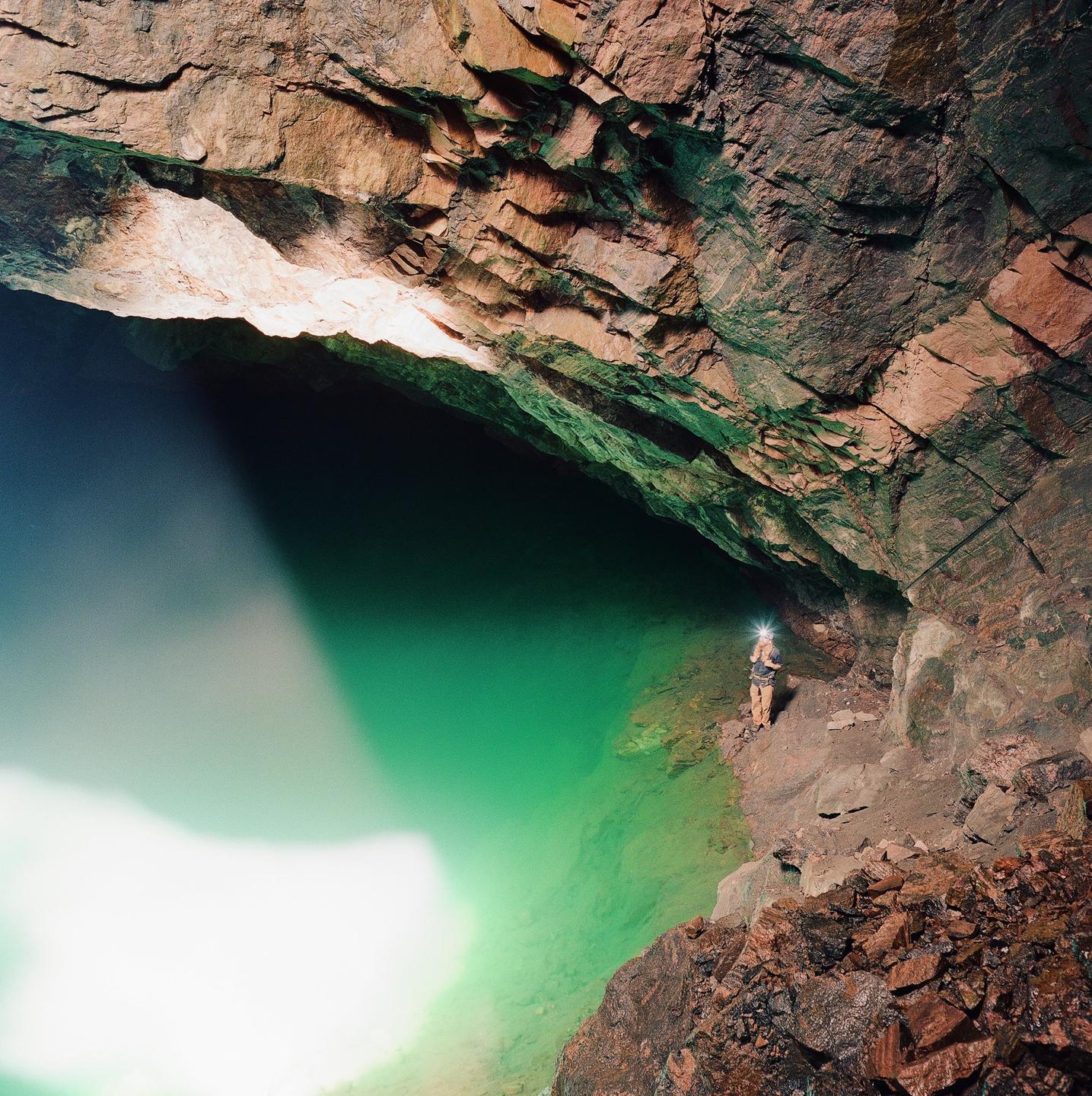 A person with a headlamp stands on a rocky ledge next to a vibrant green body of water inside a cave.