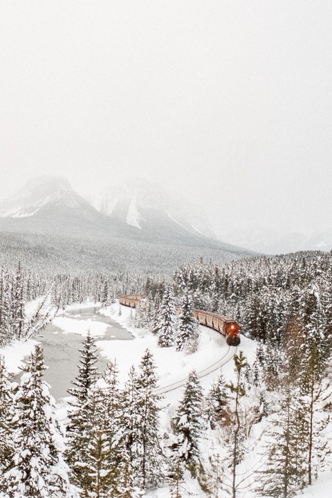 A train with an illuminated headlight travels through a snow-covered forest with mountains in the background.