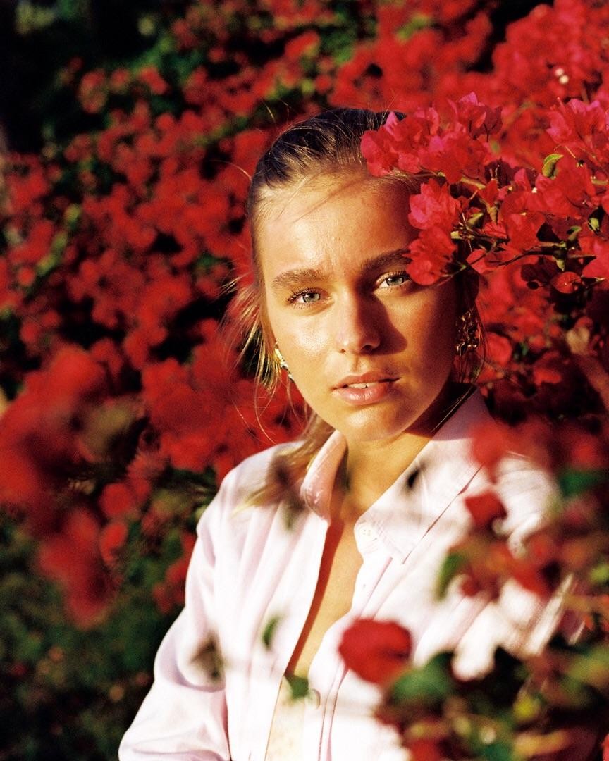 A woman with blue eyes and a pink shirt is surrounded by red flowers.