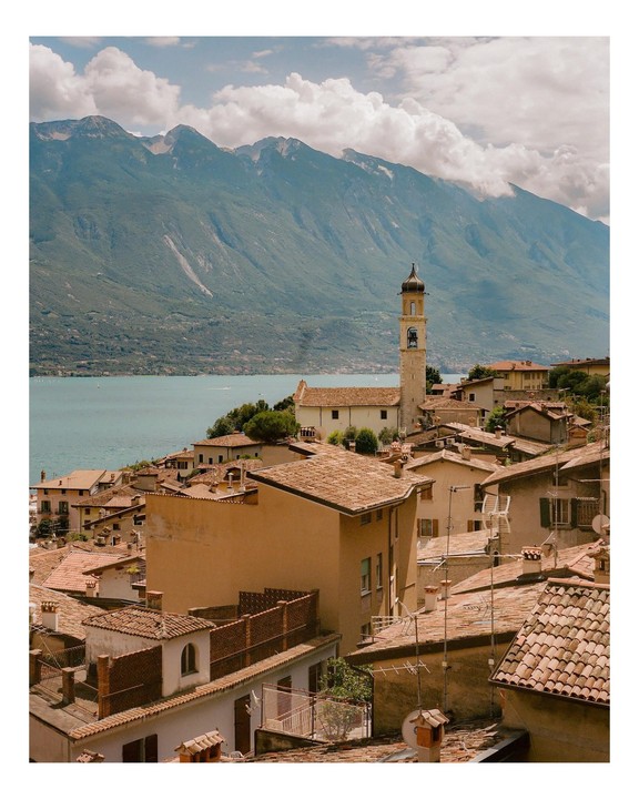 A village with a prominent bell tower sits on a hillside overlooking a large lake, with mountains rising in the background under a cloudy sky.
