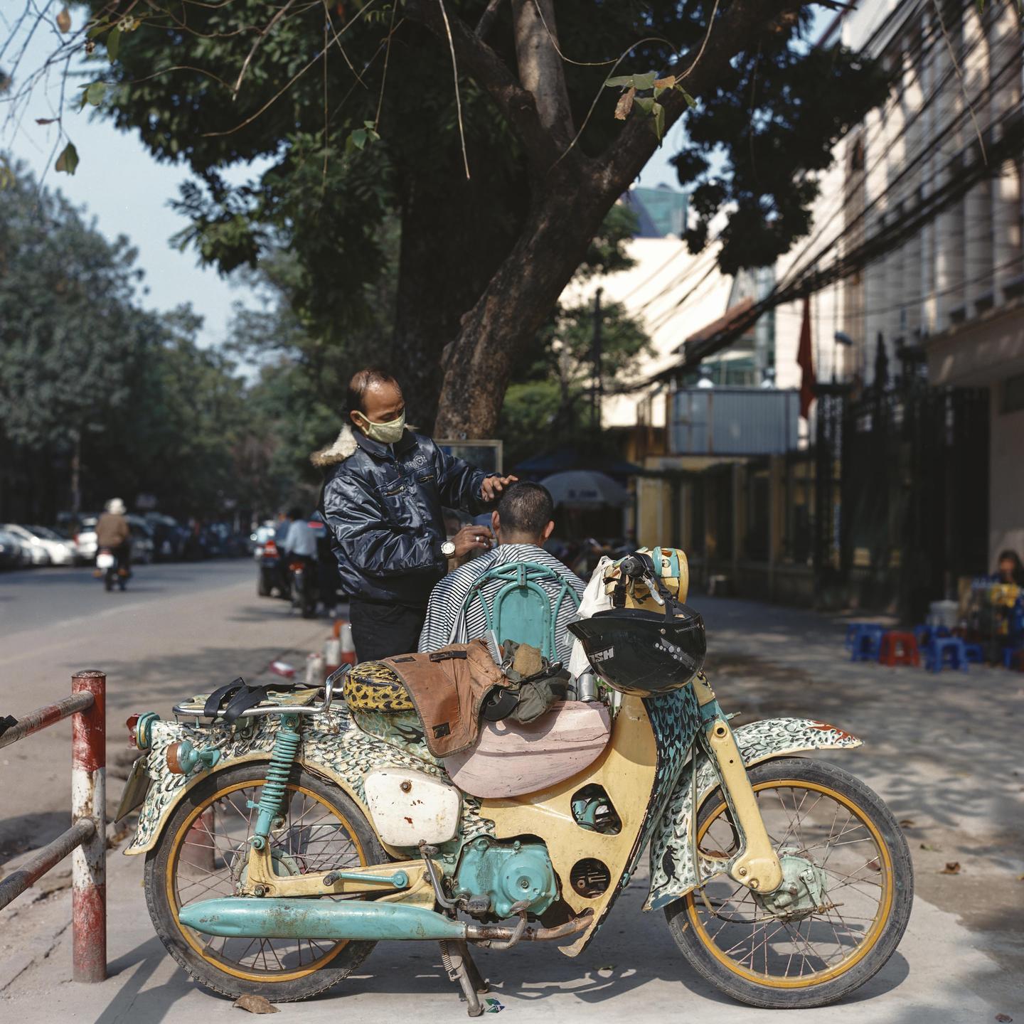 A man wearing a face mask cuts another man's hair on a street next to a colorful motorcycle.