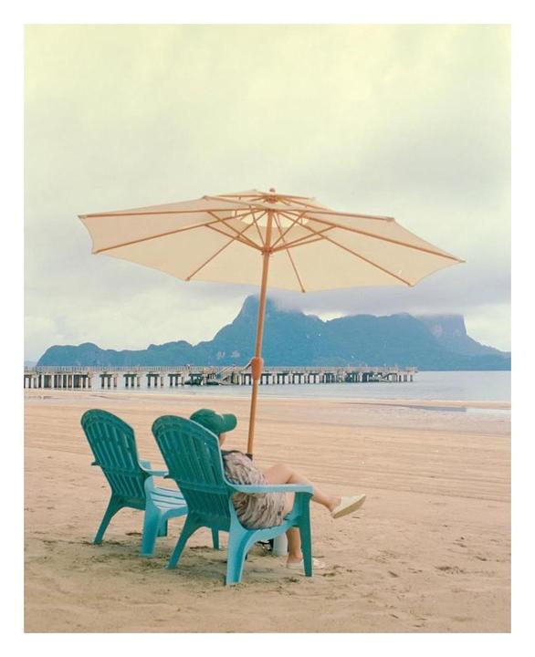 A woman sits on a beach chair under an umbrella, looking out at the ocean and mountains.
