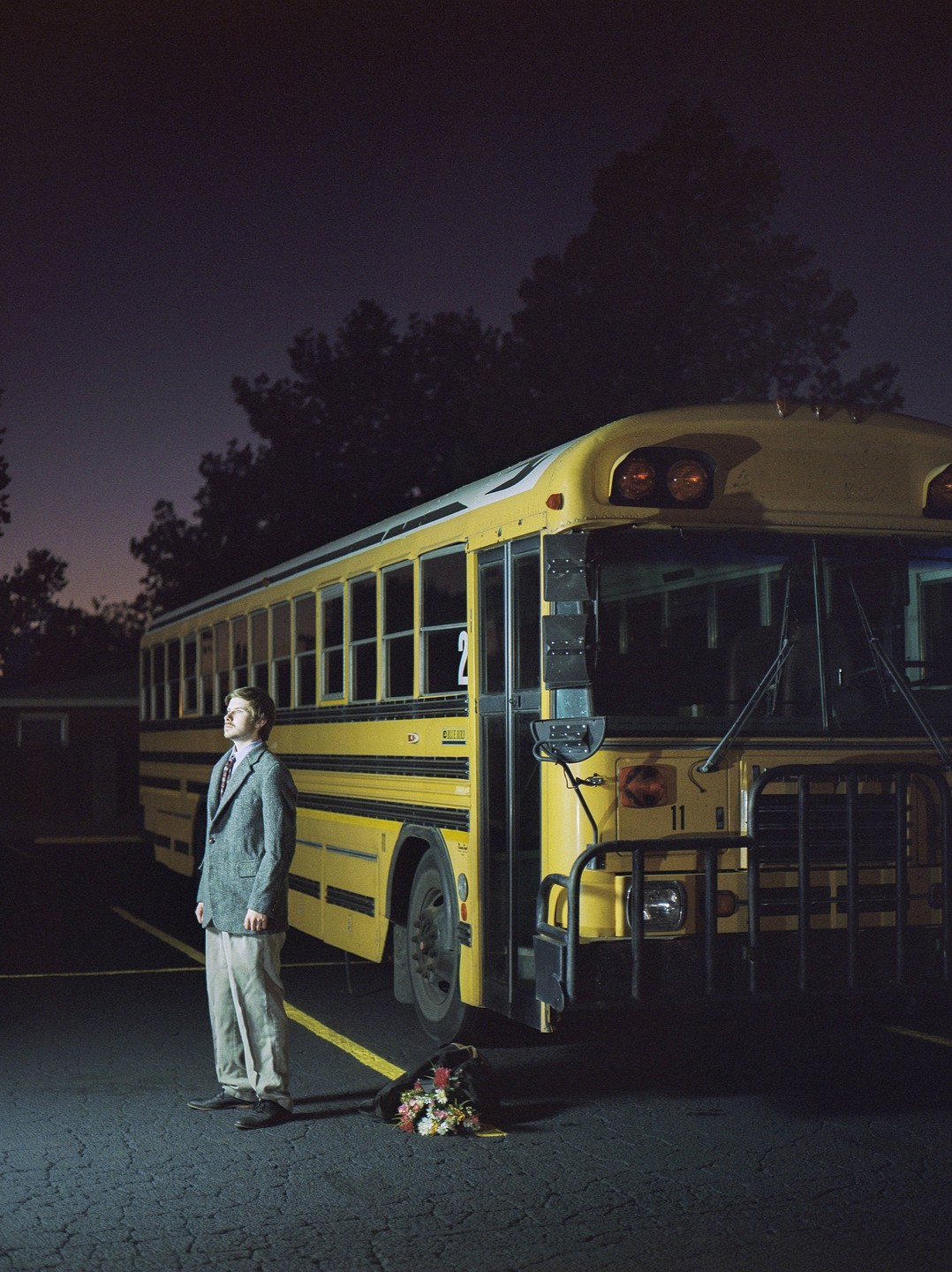 A man stands next to a yellow school bus at night, with a bouquet of flowers on the ground.