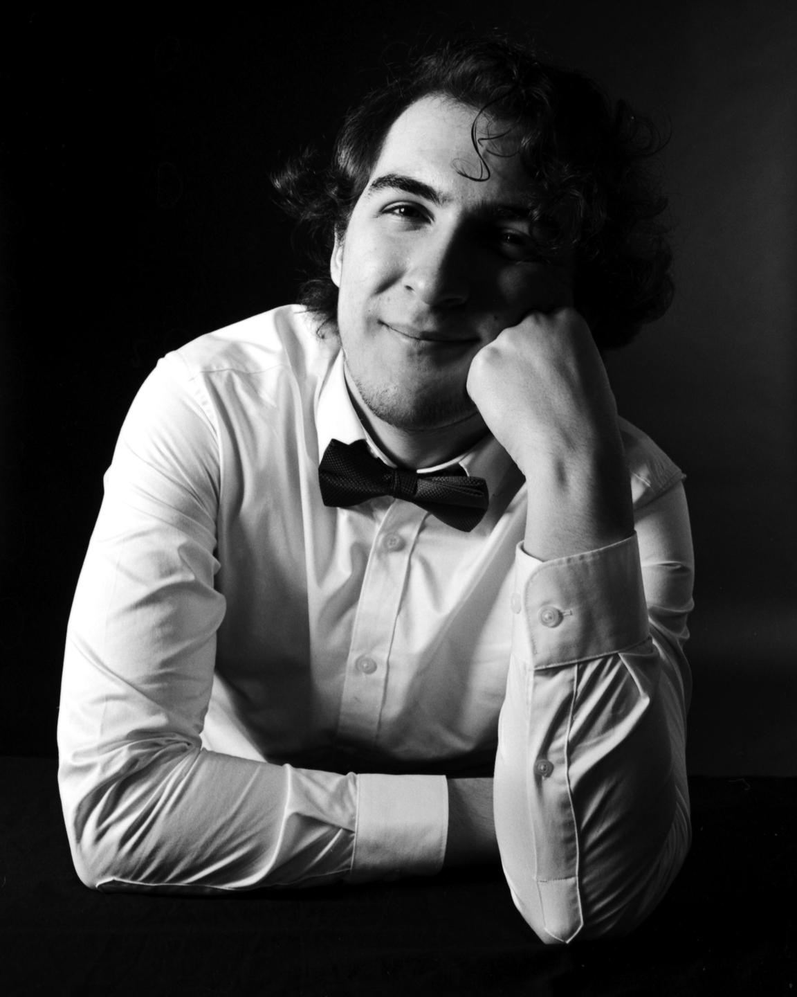 A man in a white shirt and bow tie rests his chin on his hand and smiles at the viewer.
