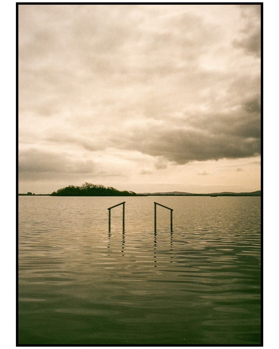 Two wooden posts emerge from the water of a lake under a cloudy sky, with a tree-covered island in the distance.