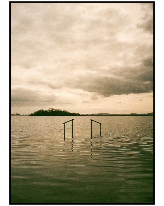 Two wooden posts emerge from the water of a lake under a cloudy sky, with a tree-covered island in the distance.