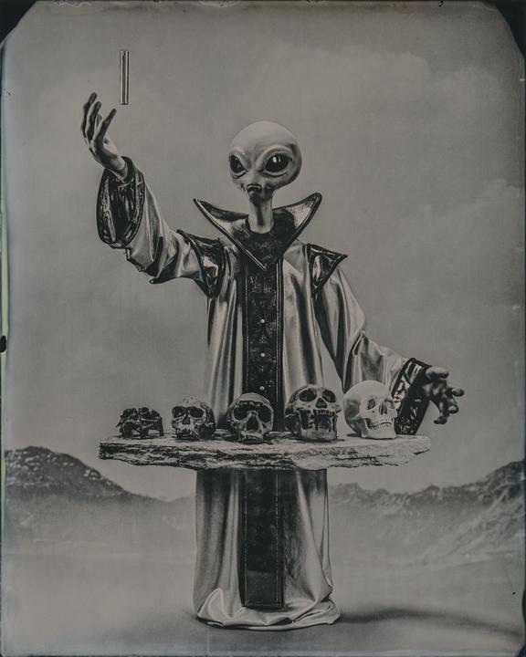 An alien in a robe holds a floating rectangular object in one hand and a tray of skulls in the other.