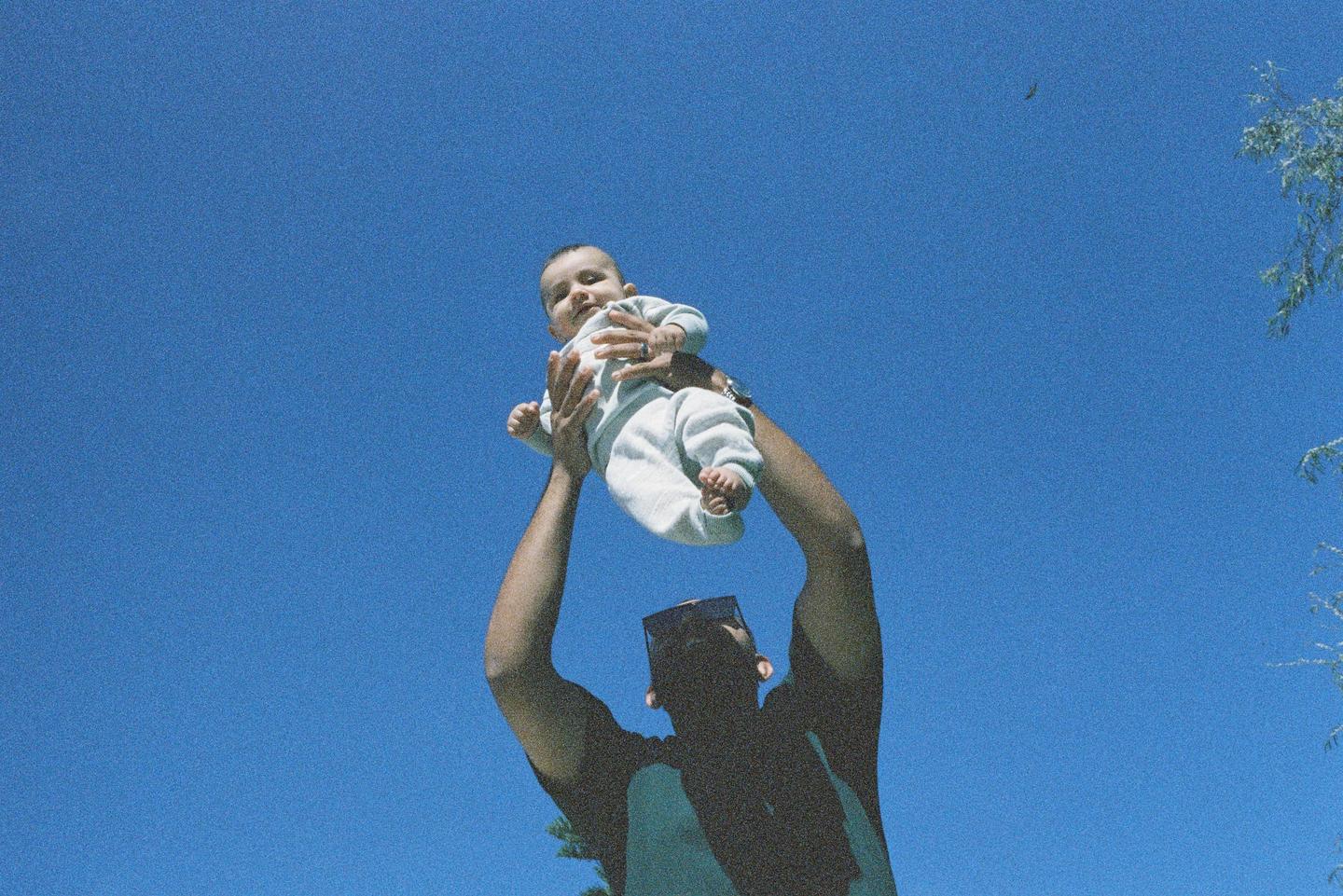 A man holds a baby up against a clear blue sky.