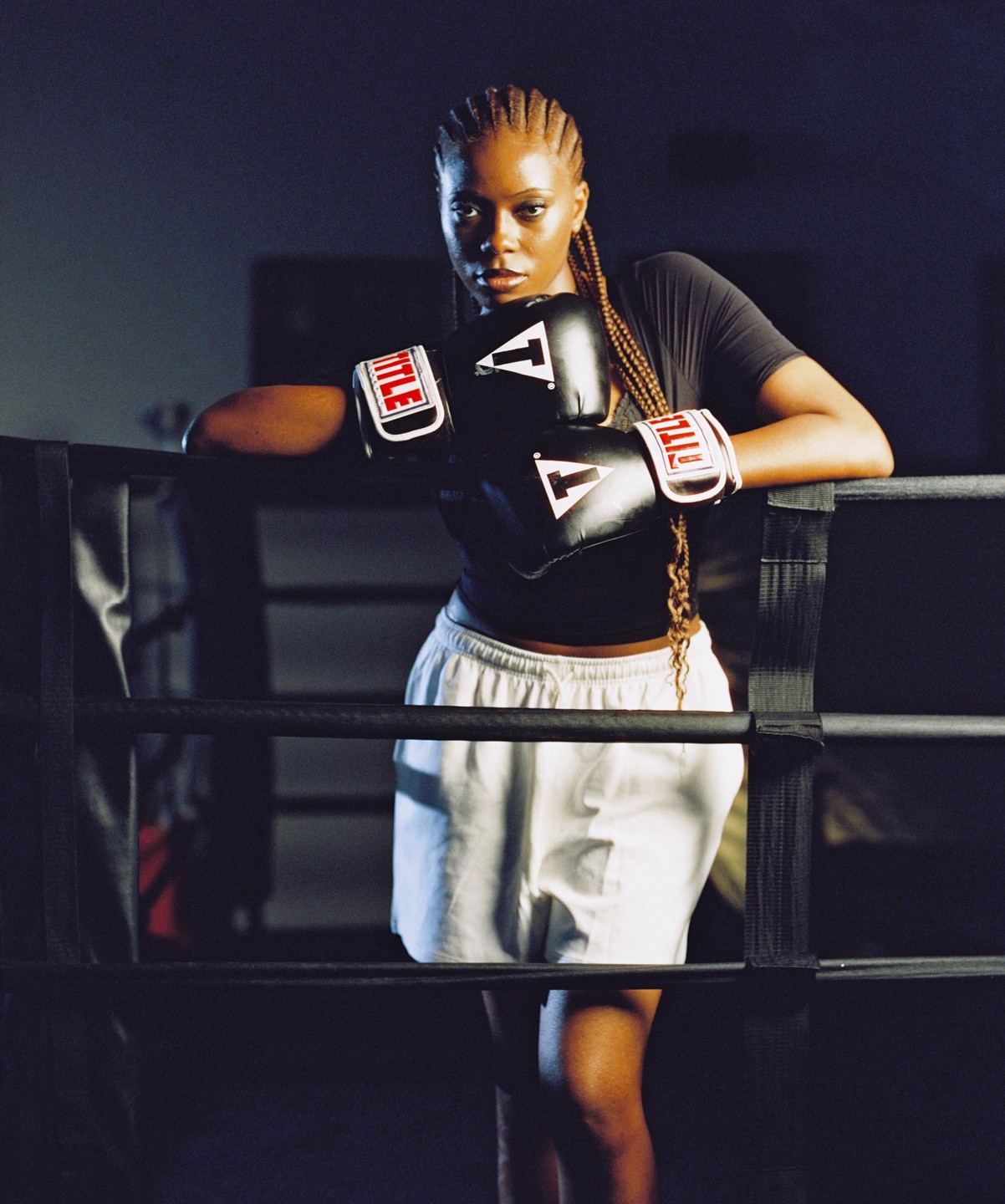 A woman wearing boxing gloves leans on the ropes of a boxing ring.