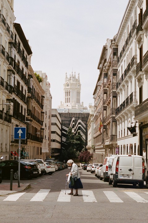 An elderly woman crosses a street in Madrid with a cane, with a large white building visible in the distance.