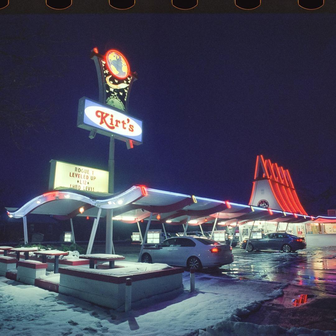 A drive-in restaurant with neon lights and cars is covered in snow at night.