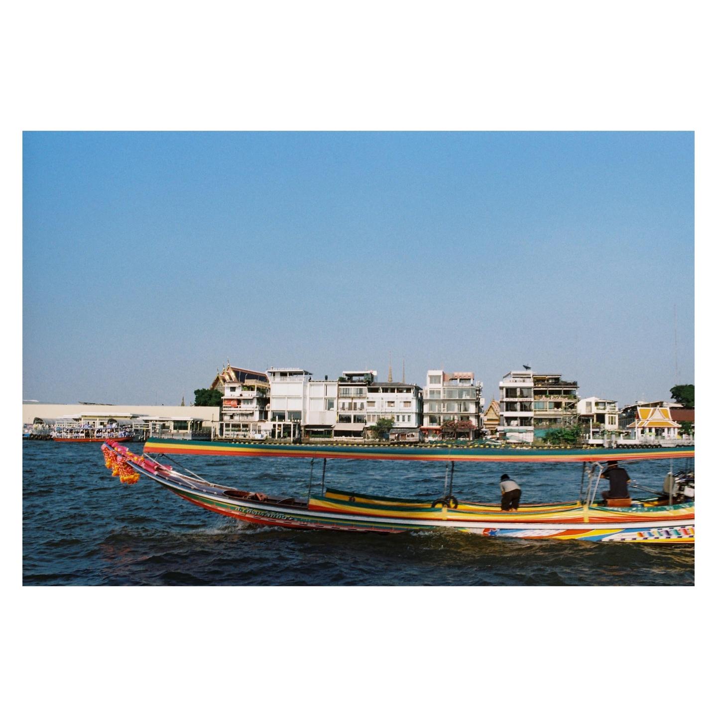 A colorful longtail boat with two men on board travels on a river with buildings on the far bank.
