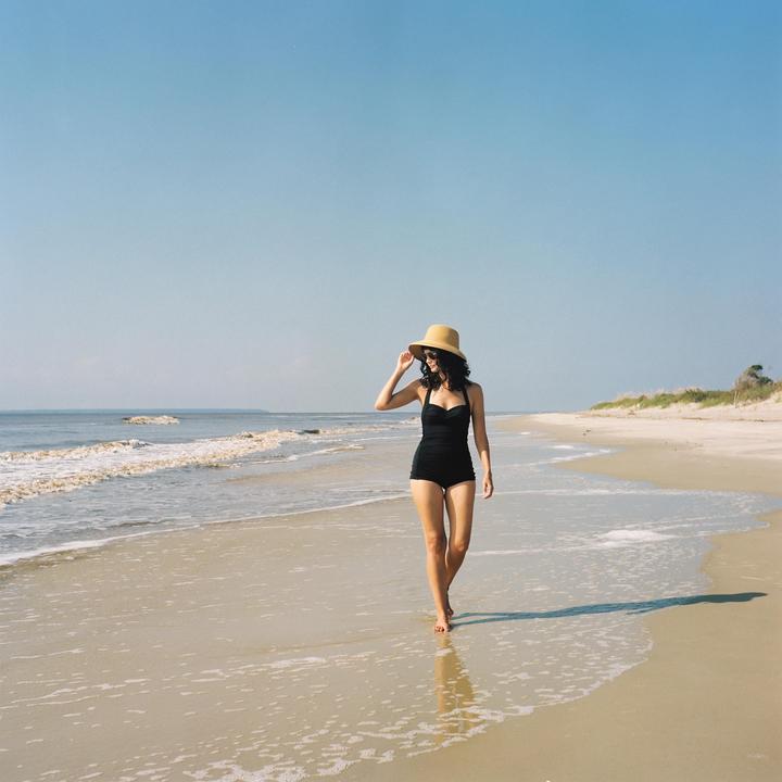 A woman in a black swimsuit and straw hat walks along a sandy beach with waves gently breaking on the shore.