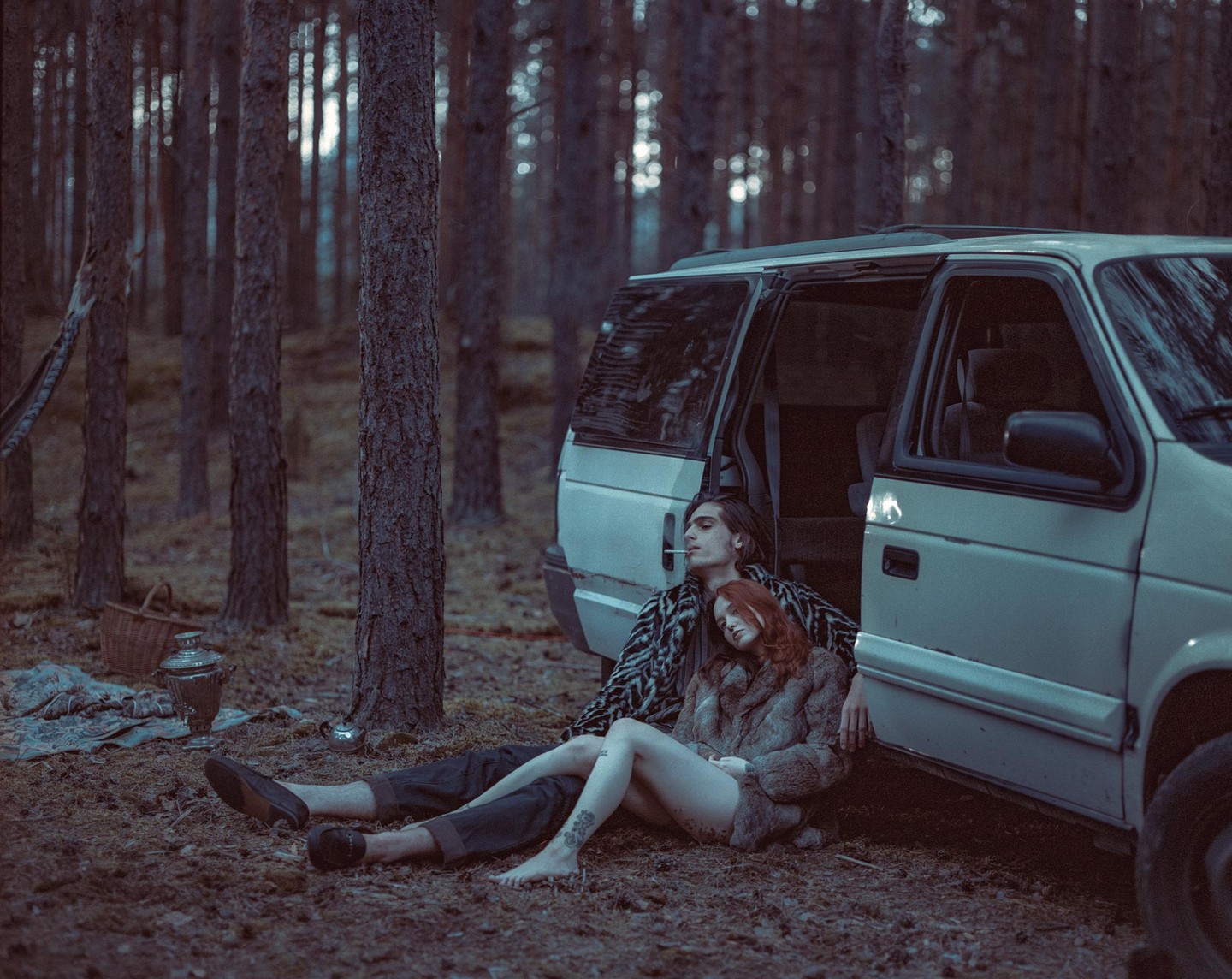 A man with a cigarette in his mouth sits with a woman resting her head on his chest, both leaning against a white van in a forest.
