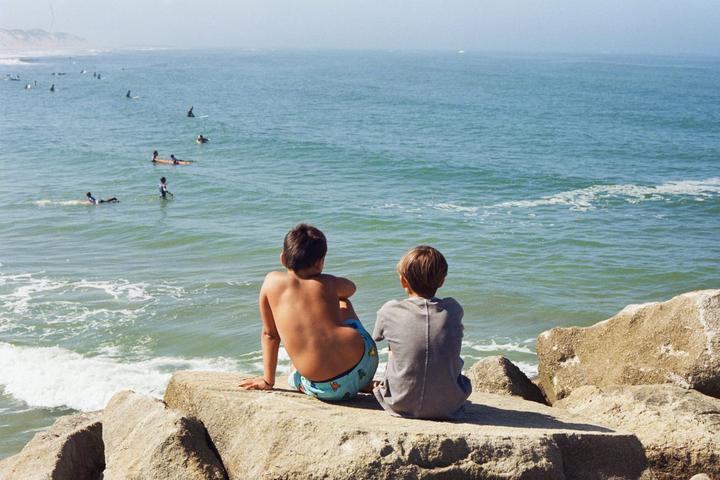Two boys sit on rocks looking out at the ocean where several people are surfing.