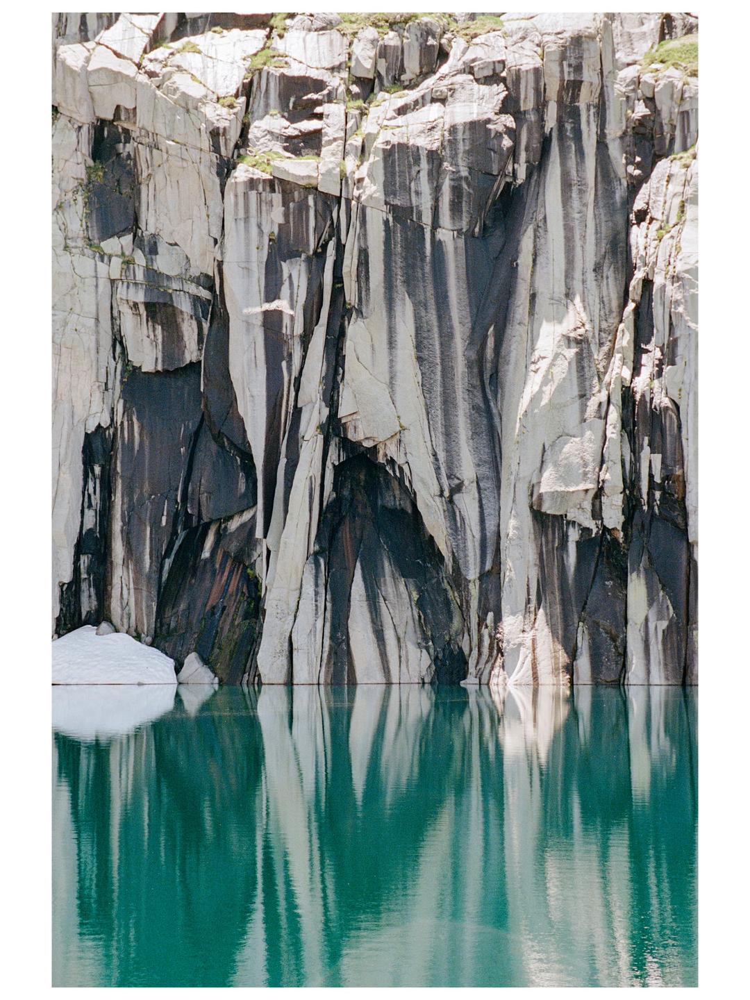 A sheer rock face with dark streaks rises from a turquoise lake, reflecting in the water.