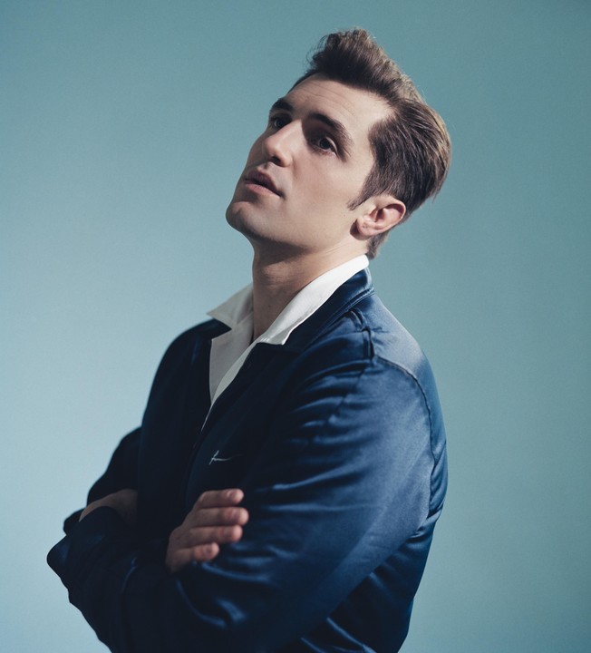 A man in a blue jacket and white shirt looks up with his arms crossed.