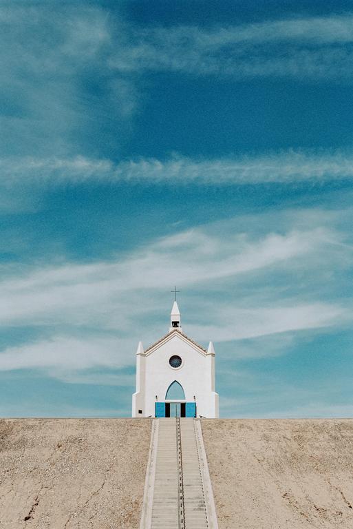 A white church with blue doors sits atop a hill, with a long staircase leading up to it under a blue sky with wispy clouds.
