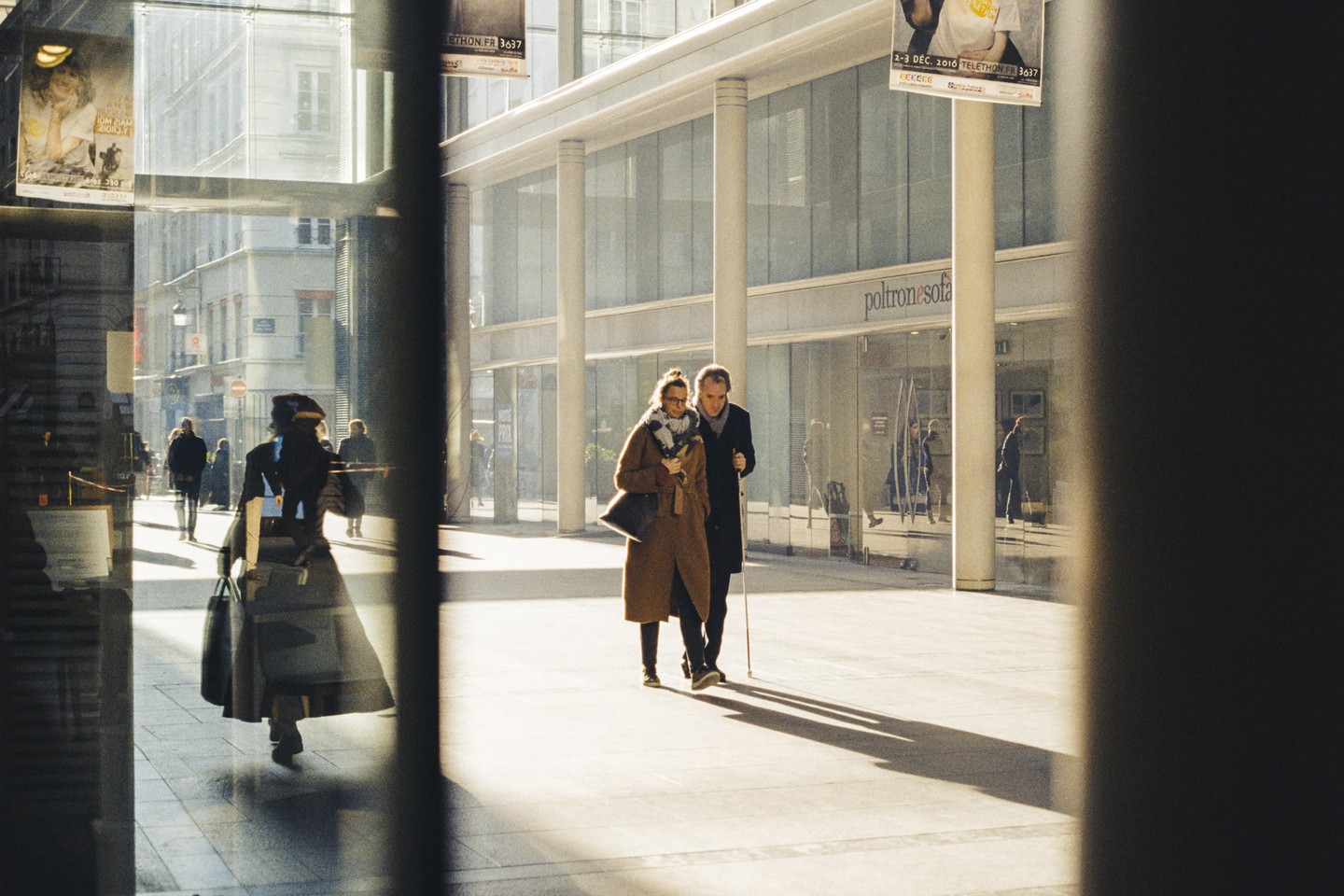 A couple walks on a sunny street with reflections in the windows of buildings.