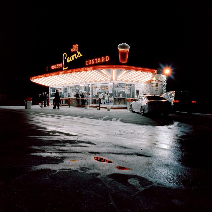 A brightly lit custard stand with neon signs stands out against the dark night, with cars and people gathered around.
