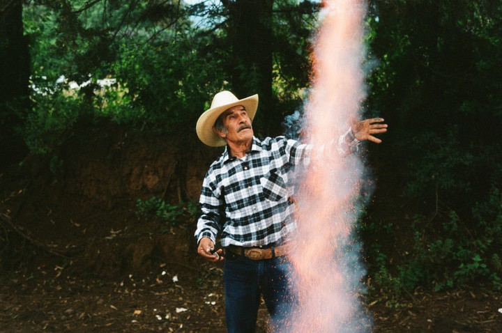 A man in a cowboy hat and plaid shirt stands next to a column of smoke and sparks.
