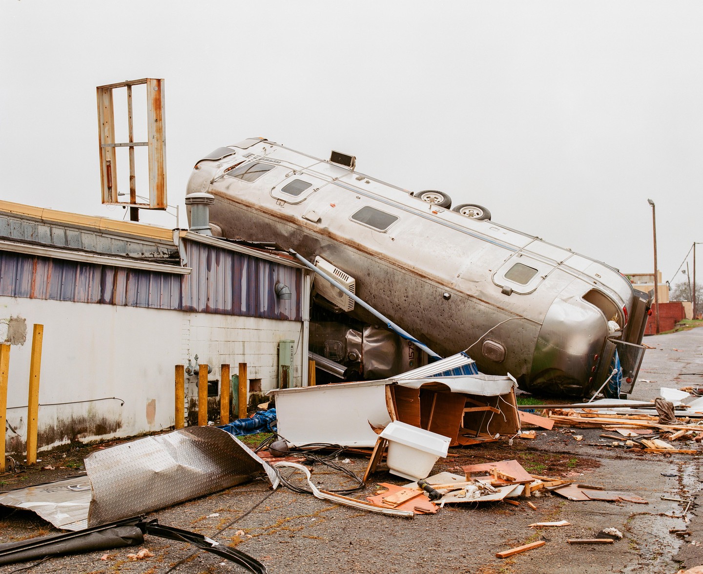 A silver trailer lies on its side, partially on top of a building, surrounded by debris.