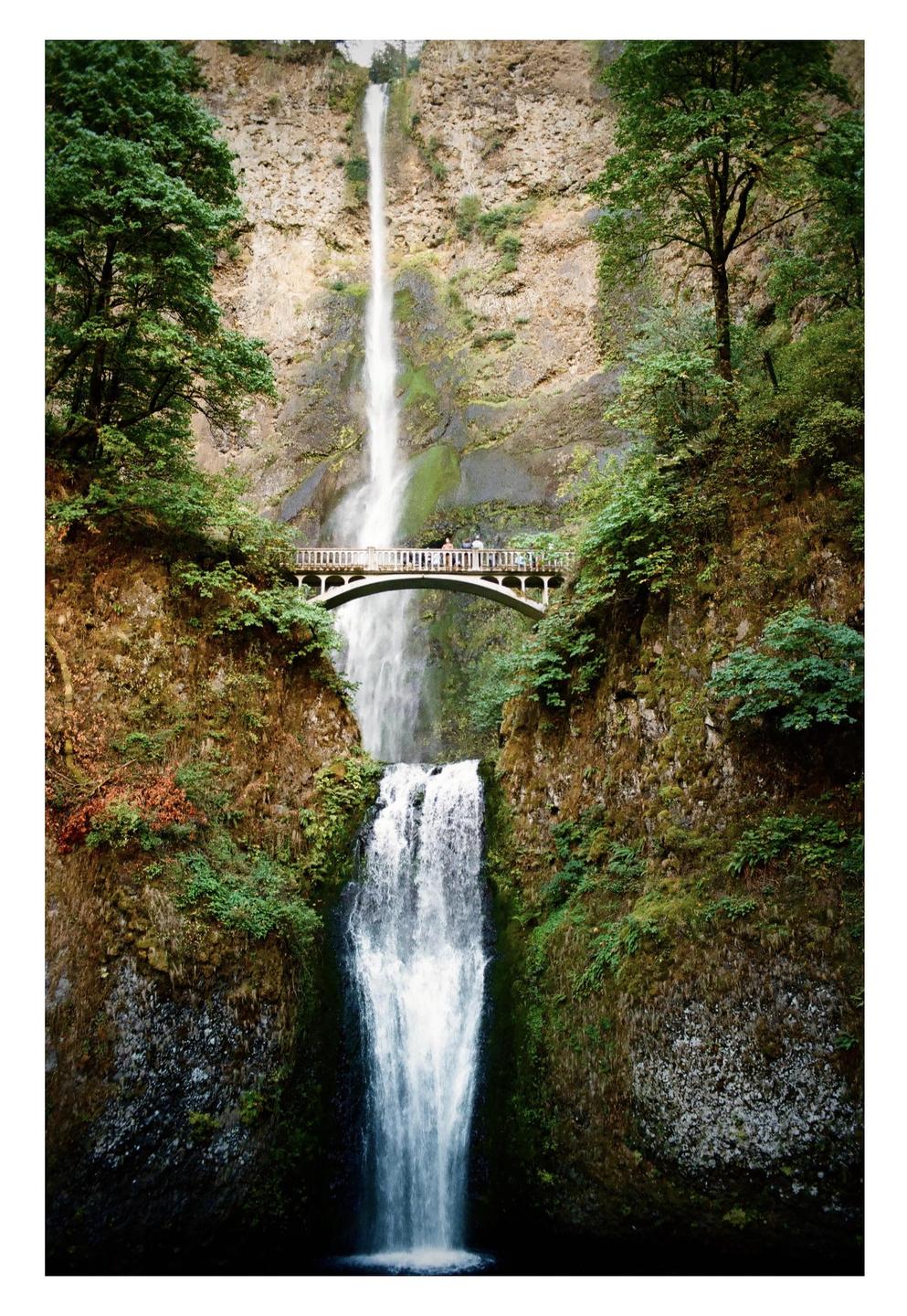 A tall waterfall cascades down a rocky cliff, with a bridge spanning the gorge in front of the upper falls.