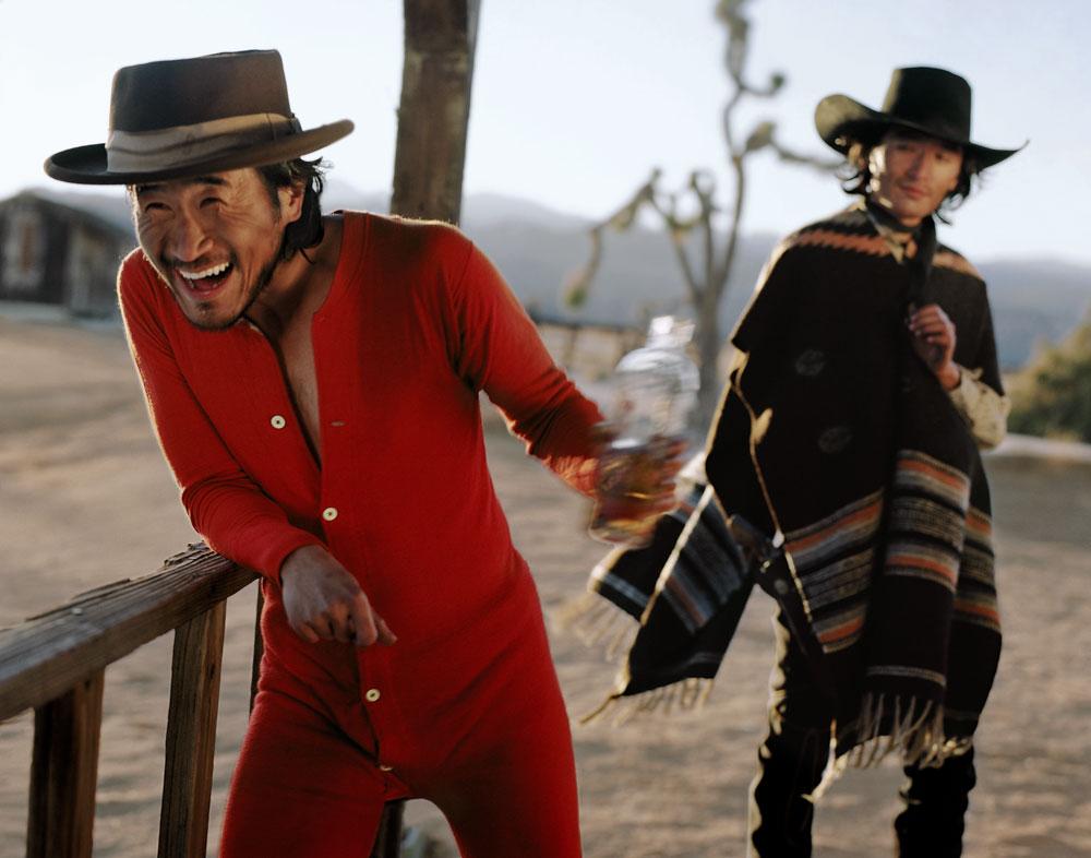 A man in a red union suit and cowboy hat laughs while holding a bottle, next to another man in a poncho and cowboy hat.