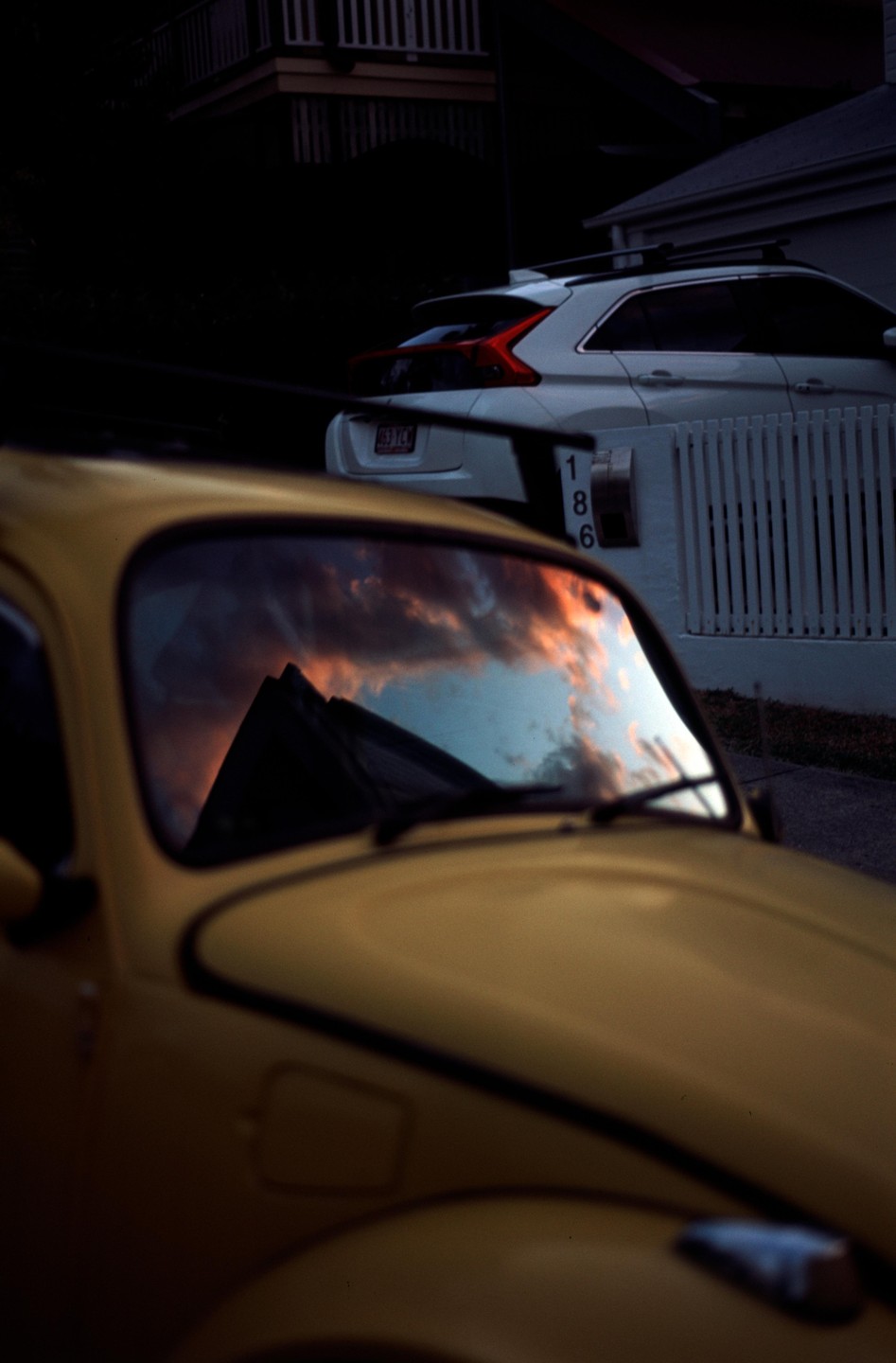 A yellow car is parked on a street with a reflection of a sunset in its windshield.